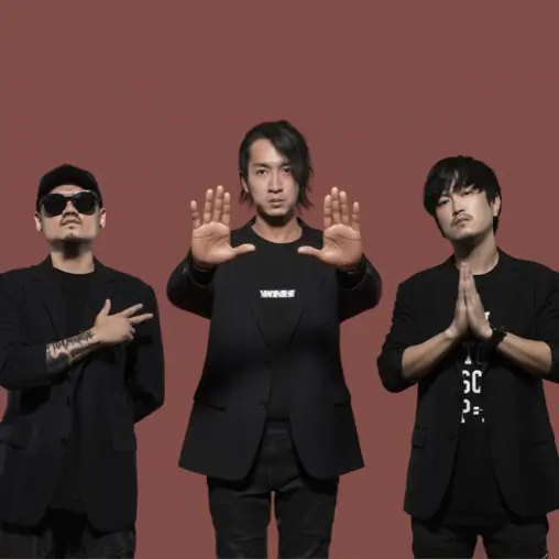 三位男團成員穿黑外套站立，中間成員雙手向前張開，左右成員各作手勢，桃紅色背景。