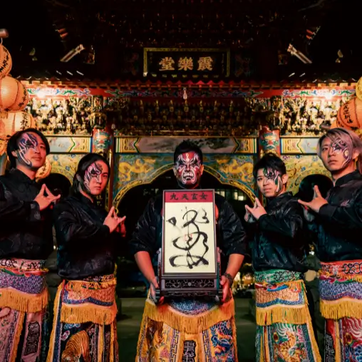 五位臉部彩繪、穿傳統陣頭服飾的表演者在寺廟牌樓前合影，中央持匾額。